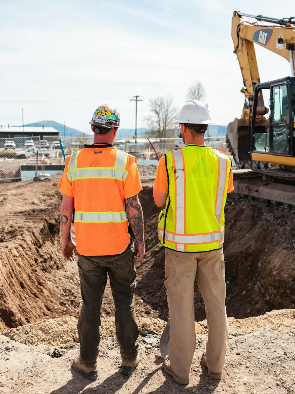 Two site workers in safety vests beside an excavator